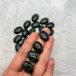 May include: A collection of oval-shaped green and black gemstones. The stones have a mottled pattern and are arranged in a pile on a white surface.