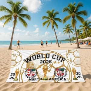 May include: A white beach towel with "WORLD CUP 2026 NORTH AMERICA" printed in gold and black. The towel features soccer balls, trophy illustrations, and flags. Palm trees and people playing soccer are in the background.