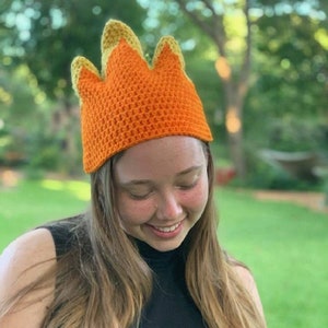 May include: A crocheted crown hat with orange and yellow flames. The hat is worn by a person with long brown hair.