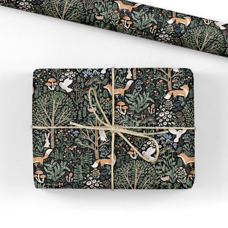 May include: A rectangular gift wrapped in black paper featuring a forest pattern with orange foxes, white rabbits, owls, mushrooms, and green trees, tied with tan twine. A matching roll of wrapping paper is positioned above the gift.