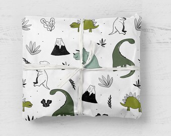 May include: A square package wrapped in white paper featuring a repeating pattern of green and black line-art dinosaurs, volcanoes, and foliage. The paper is secured with thin white string tied in a simple cross pattern across the centre.