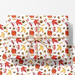 May include: A rectangular gift wrapped in white paper featuring a repeating watercolour pattern of red maple leaves, yellow foliage, brown acorns, and red berries, tied with a natural tan jute twine bow centred on the front surface.