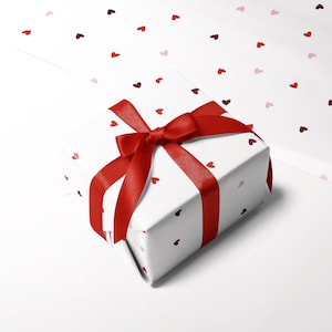 May include: A white gift box with a red ribbon and bow. The box is wrapped in paper with a pattern of small red hearts.