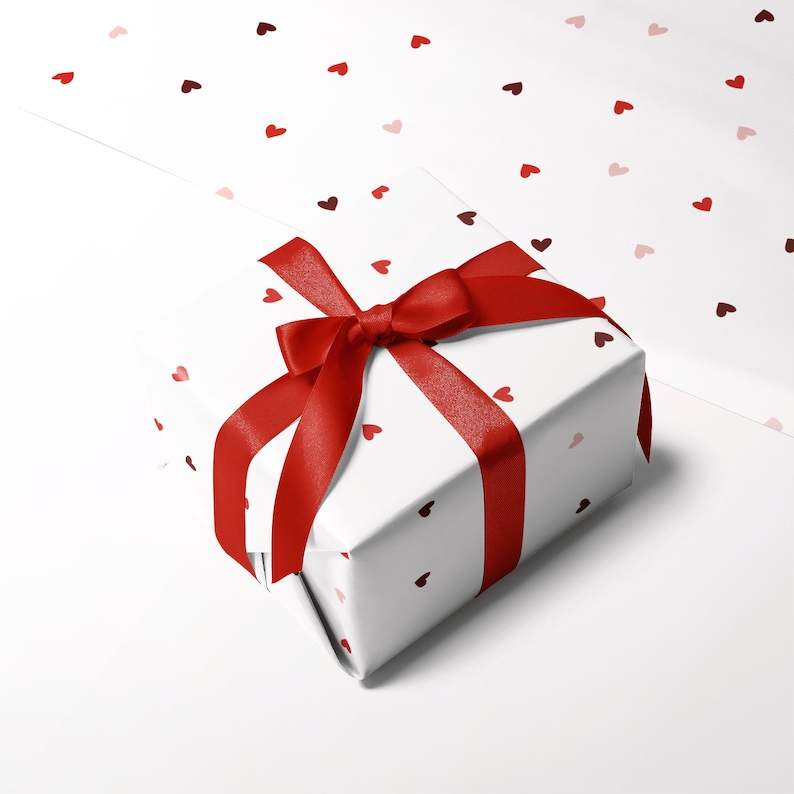 May include: A white gift box with a red ribbon and bow. The box is wrapped in paper with a pattern of small red hearts.