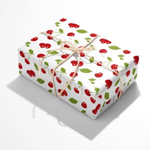 Peut inclure: Une bo&icirc;te cadeau rectangulaire envelopp&eacute;e dans du papier blanc avec un motif r&eacute;p&eacute;titif de cerises rouges accompagn&eacute;es de feuilles et de tiges vertes. Le paquet est nou&eacute; avec une fine ficelle de jute naturelle formant un n&oelig;ud sur le dessus.