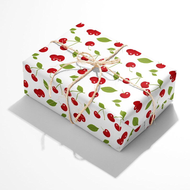 Peut inclure: Une bo&icirc;te cadeau rectangulaire envelopp&eacute;e dans du papier blanc avec un motif r&eacute;p&eacute;titif de cerises rouges accompagn&eacute;es de feuilles et de tiges vertes. Le paquet est nou&eacute; avec une fine ficelle de jute naturelle formant un n&oelig;ud sur le dessus.