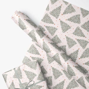 May include: Wrapping paper featuring a repeating pattern of sage green pine trees and small copper-colored stars on a pale pink background. The paper is shown in a roll and also wrapped around a rectangular gift box with a sage green ribbon.