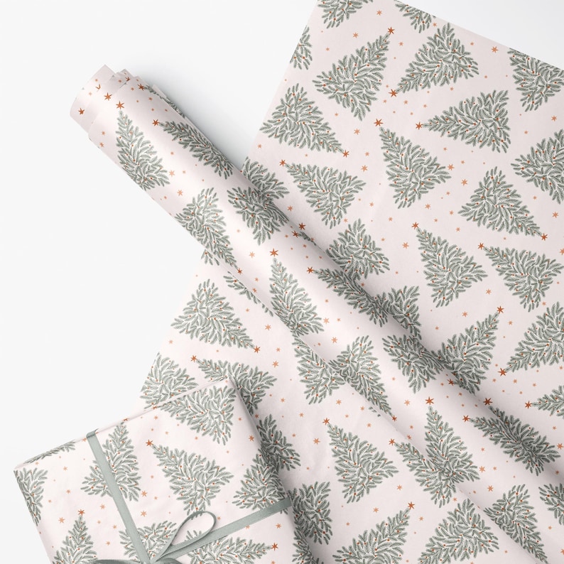 May include: Wrapping paper featuring a repeating pattern of sage green pine trees and small copper-colored stars on a pale pink background. The paper is shown in a roll and also wrapped around a rectangular gift box with a sage green ribbon.