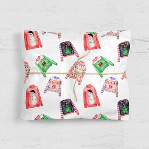 May include: White wrapping paper featuring a repeating watercolor pattern of various holiday sweaters, including red snowman designs, green sweaters with gift boxes, dark blue tree patterns, and pink sweaters with colorful string light graphics.