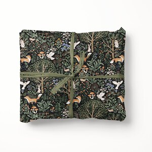 May include: A rectangular package wrapped in black paper featuring a repeating forest pattern of orange foxes, white owls, rabbits, mushrooms, and green trees, tied with a thin olive green ribbon centred in a cross pattern across the surface.