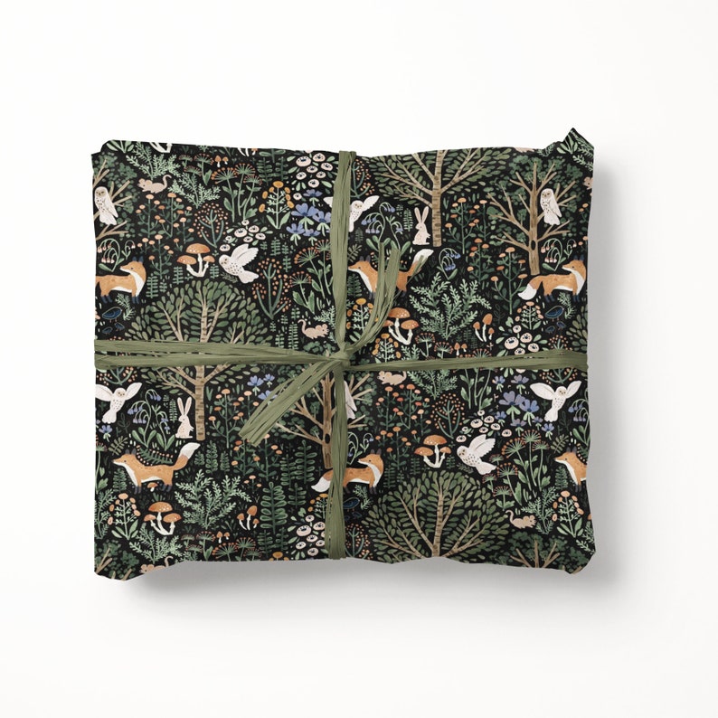 May include: A rectangular package wrapped in black paper featuring a repeating forest pattern of orange foxes, white owls, rabbits, mushrooms, and green trees, tied with a thin olive green ribbon centred in a cross pattern across the surface.