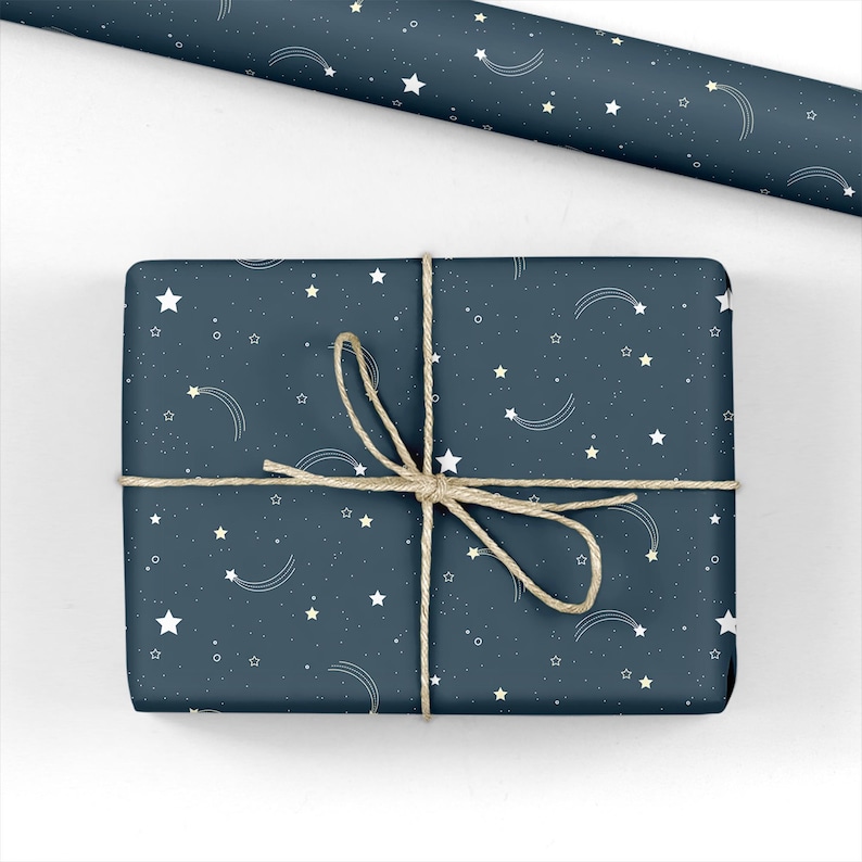 May include: A wrapped gift with a navy blue background, adorned with white stars and shooting star designs. The gift is tied with natural twine. A roll of matching wrapping paper is visible in the background.