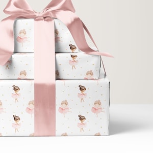 May include: Three stacked rectangular gift boxes wrapped in white paper featuring a repeating pattern of small ballerinas in pink tutus and tiny gold stars. A wide, smooth, light pink satin ribbon is tied in a large bow on the top box.