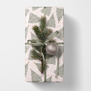 May include: A rectangular gift box wrapped in white paper featuring a repeating pattern of sage green pine trees and small orange stars. The package is tied with a sage green ribbon, a sprig of evergreen needles, and a silver glitter ornament.