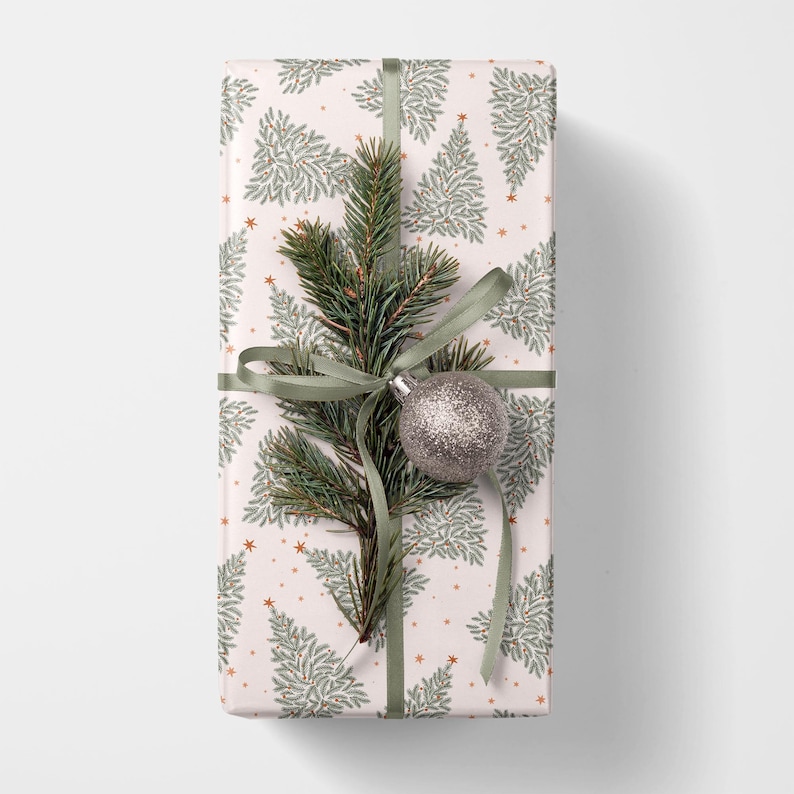 May include: A rectangular gift box wrapped in white paper featuring a repeating pattern of sage green pine trees and small orange stars. The package is tied with a sage green ribbon, a sprig of evergreen needles, and a silver glitter ornament.