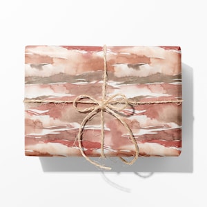May include: A rectangular gift box wrapped in paper featuring a horizontal watercolour pattern in shades of terracotta, dusty rose, and muted brown. The package is tied with a natural tan jute twine string arranged in a simple bow at the centre.