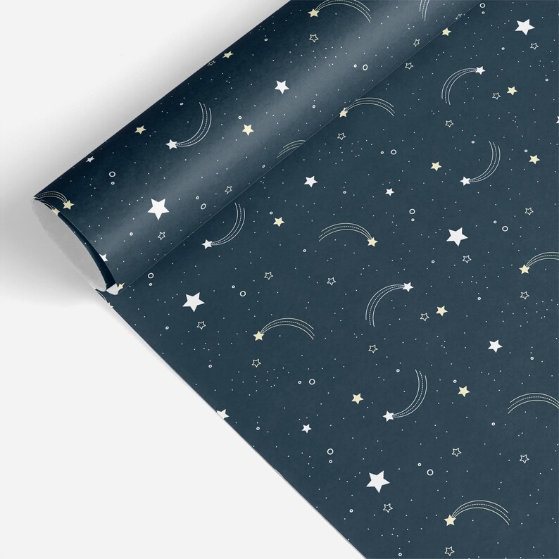 May include: A roll of navy blue wrapping paper with a celestial pattern. The design features white stars, shooting stars, and small dots scattered across the surface. The paper is partially unrolled, revealing the pattern.