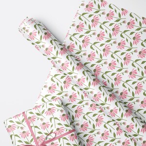 May include: A roll of wrapping paper and a gift-wrapped box featuring a repeating pattern of pink coneflower blossoms with dark brown centers and green stems on a white background. The gift is tied with a thin pink ribbon.