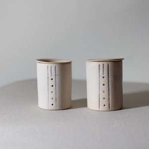Op de afbeelding: Twee crèmekleurige keramische potten met deksels. Elke cilindrische container heeft een reeks verticale zwarte lijnen en kleine, gelijkmatig verdeelde stippen. De deksels hebben een bijpassende crèmekleur. De potten staan tegen een neutrale achtergrond.