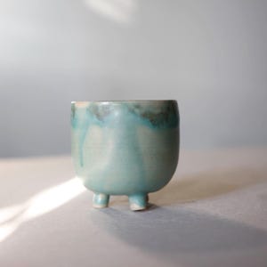 May include: A small, turquoise ceramic bowl with three short legs. The bowl has a rounded shape and a glossy finish with darker blue and brown accents around the rim. The glaze has a dripped effect, adding visual interest.