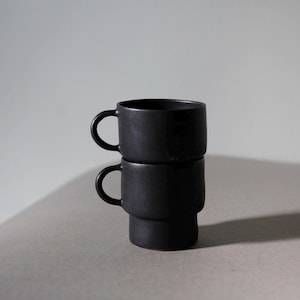 Può includere: Due tazze in ceramica nera opaca impilate l'una sull'altra. Ogni tazza ha un manico arrotondato e un corpo cilindrico. Le tazze sono su uno sfondo neutro, con un'illuminazione soffusa. Misurano circa 8 cm di altezza.