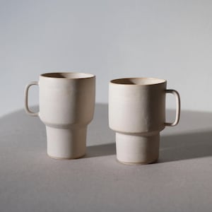May include: Two off-white ceramic mugs with a stepped design. Each mug has a cylindrical body, a handle, and a slightly flared rim. The mugs are displayed on a neutral surface, with soft lighting.