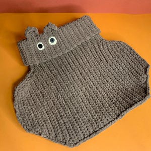 Puede incluir: Una manta de bebé de ganchillo gris hecha a mano con forma de hipopótamo. La manta presenta una cabeza con orejas pequeñas y dos ojos de botón. La manta está sobre un fondo naranja.