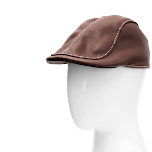 Könnte beinhalten: Braune Leder-Flatcap mit weißen Nähten. Die Kappe wird auf einem weißen Schaufensterkopf präsentiert. Der Hut hat einen gebogenen Schirm und eine strukturierte Oberfläche.