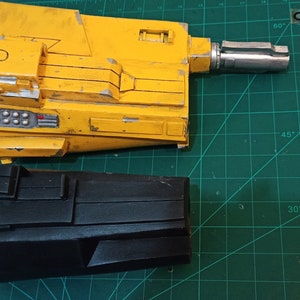 May include: Two toy gun parts, one yellow and one black. The yellow part has a silver barrel and a series of small, round, silver details. The black part has a series of rectangular details.