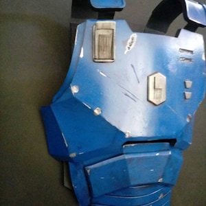 May include: A blue and gray plastic chest piece with a textured surface and a silver panel. The chest piece has a worn look with scratches and scuffs.