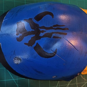 May include: A blue and yellow helmet with a black Mandalorian symbol painted on the front. The helmet has a black trim around the edge.