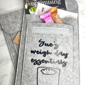 May include: A gray felt organizer with the text "Sue's weight day essentials" and a scale graphic. The organizer contains a magazine, brochures, and two snack bars. The magazine cover reads "food optimising".