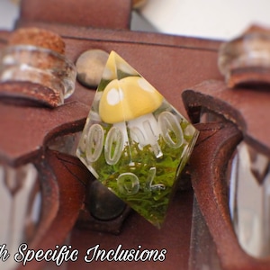 May include: A clear resin dice with a yellow mushroom and green moss inside. The dice is shaped like a pyramid and has the numbers 1-6 on its sides. The dice is sitting on a brown leather pouch with two small glass vials.