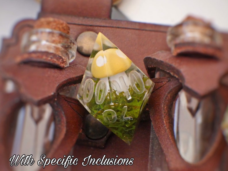 May include: A clear resin dice with a yellow mushroom and green moss inside. The dice is shaped like a pyramid and has the numbers 1-6 on its sides. The dice is sitting on a brown leather pouch with two small glass vials.