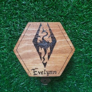 May include: A hexagonal wooden box featuring the Skyrim logo, etched in dark lines, and the name "Evelynn" beneath. The box has a keyhole and sits on a green, grassy surface. The wood grain is visible.