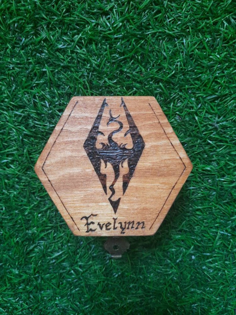 May include: A hexagonal wooden box featuring the Skyrim logo, etched in dark lines, and the name "Evelynn" beneath. The box has a keyhole and sits on a green, grassy surface. The wood grain is visible.