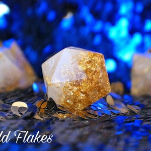 May include: A white, translucent, six-sided dice with gold flakes embedded in the resin. The dice is sitting on a black surface with blue sequins.