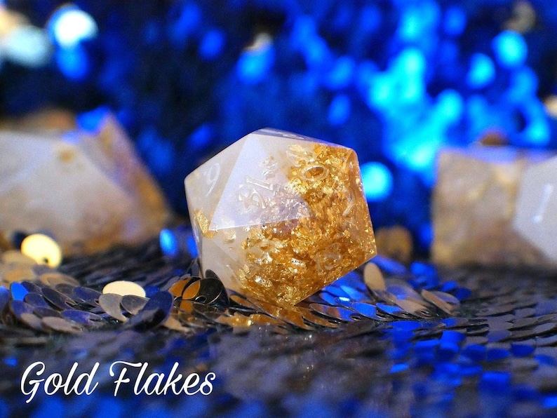 May include: A white, translucent, six-sided dice with gold flakes embedded in the resin. The dice is sitting on a black surface with blue sequins.