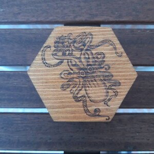 May include: Hexagonal wooden coaster featuring an etched octopus design. The octopus is centrally placed, with its tentacles spreading out. The wood has a natural grain and a warm, light brown colour.