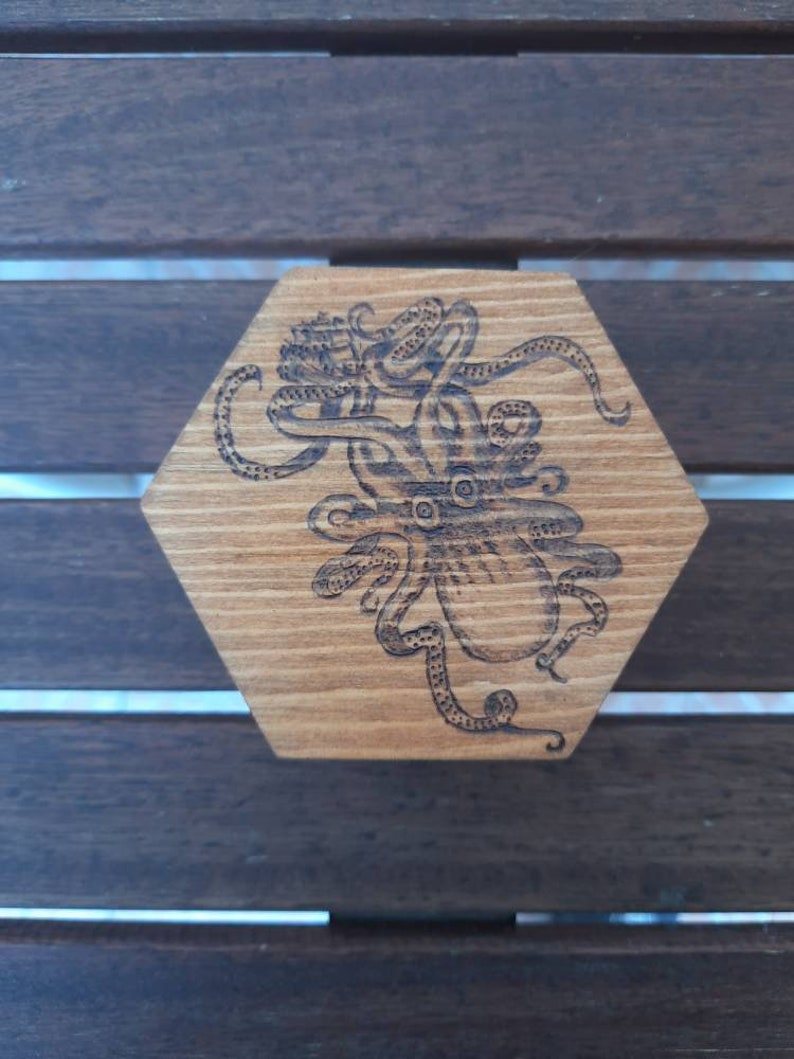 May include: Hexagonal wooden coaster featuring an etched octopus design. The octopus is centrally placed, with its tentacles spreading out. The wood has a natural grain and a warm, light brown colour.