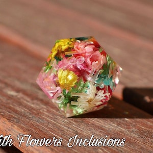 May include: A multi-sided, clear resin dice with a variety of pressed flowers and leaves inside. The dice is a light pink, yellow, and green color.