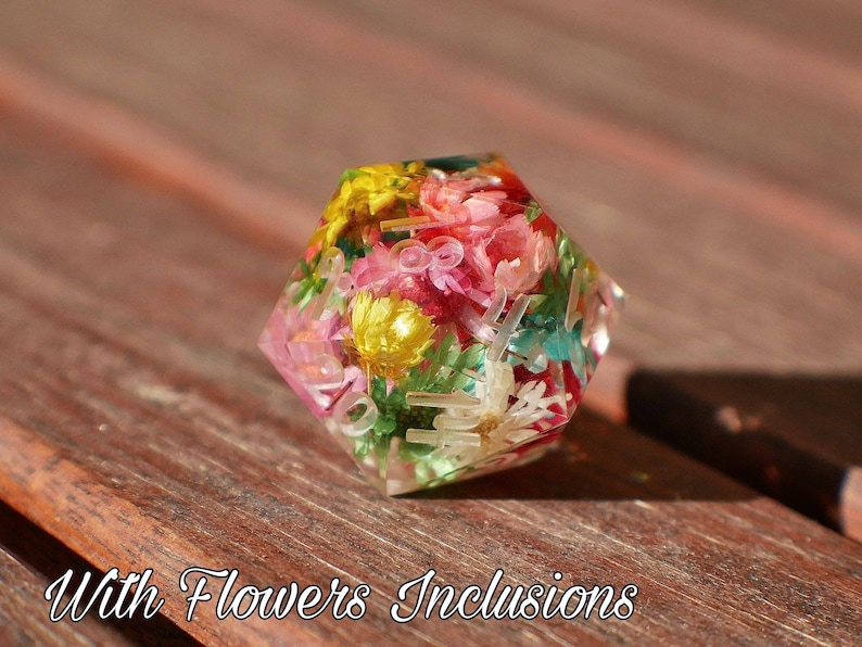 May include: A multi-sided, clear resin dice with a variety of pressed flowers and leaves inside. The dice is a light pink, yellow, and green color.