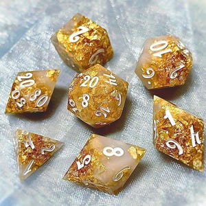 May include: A set of seven polyhedral dice with a gold leaf and cream resin design. Each die features white numbering. The dice are designed for tabletop role-playing games.