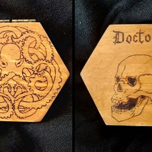 May include: Hexagonal wooden box with a hinged lid. One side features an etched octopus design surrounded by gears. The other side has the word "Doctor" above a skull illustration. The box is made of light-coloured wood.
