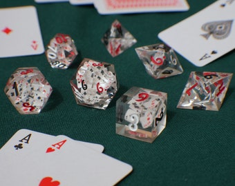 May include: Clear resin dice with playing cards embedded inside, featuring red and white numbers. The dice are scattered on a green felt surface, with playing cards visible in the background.
