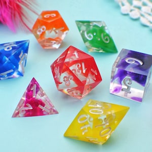 May include: A set of six colourful dice with clear plastic bodies and white numbers. The dice are in various shapes, including a pyramid, a cube and a diamond. The dice are on a light blue surface.