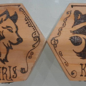 May include: Two hexagonal wooden plaques with pyrography designs. One features a wolf, the other a raccoon. Each plaque has a name, "CHRIS" and "KRIS", and decorative borders. The wood has a light brown colour.