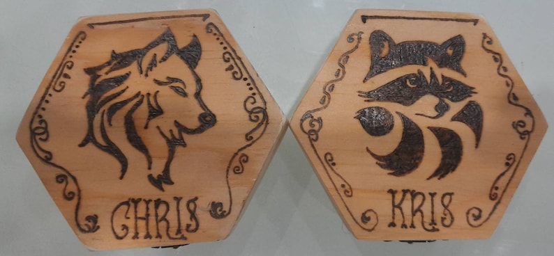 May include: Two hexagonal wooden plaques with pyrography designs. One features a wolf, the other a raccoon. Each plaque has a name, "CHRIS" and "KRIS", and decorative borders. The wood has a light brown colour.