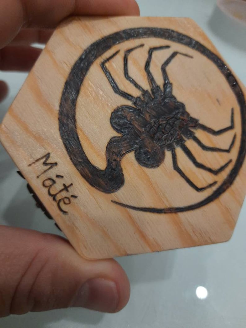 May include: Hexagonal wooden coaster featuring a dark brown insect design within a black circle. The word "Mat&eacute;" is written in black on the side. The coaster is made of light-coloured wood, with a natural grain.