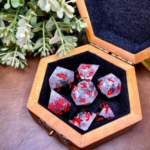 May include: A set of seven clear resin dice with red numbering, presented in a wooden hexagonal box lined with black felt. The dice have a marbled effect, suitable for tabletop games.